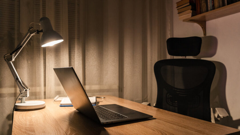 Open laptop on a wooden desk lit by an angled desk lamp in the evening, with an empty office chair beside it