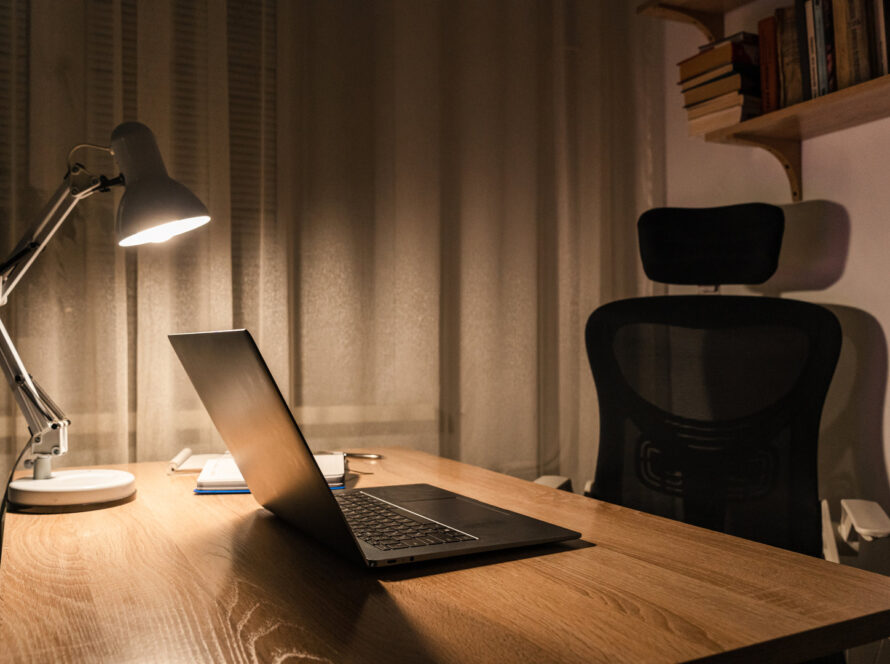 Open laptop on a wooden desk lit by an angled desk lamp in the evening, with an empty office chair beside it