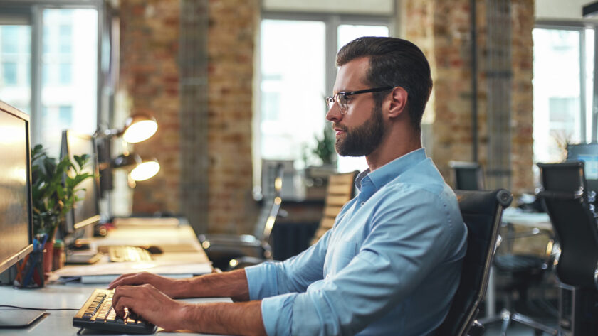 Man in a light blue shirt typing at a keyboard at a desk in a bright open plan office with brick walls and large windows