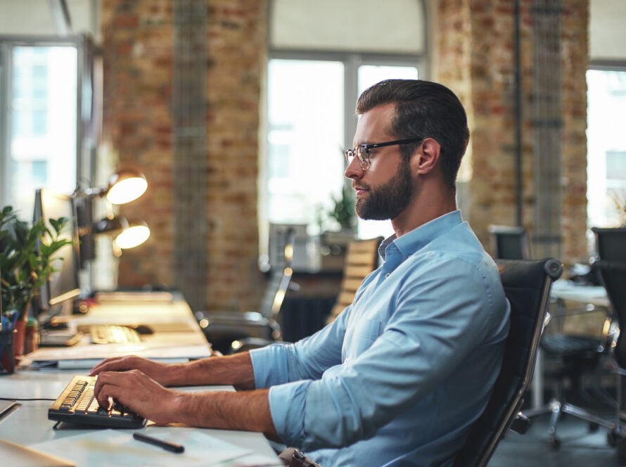 Man in a light blue shirt typing at a keyboard at a desk in a bright open plan office with brick walls and large windows