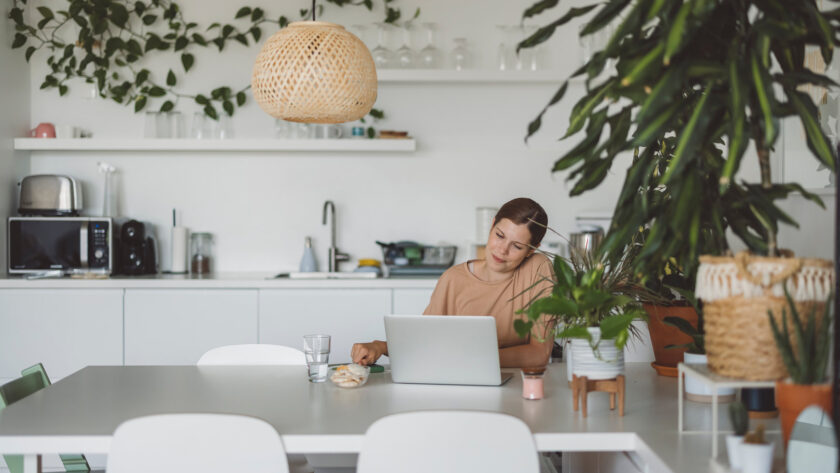 Woman in a tan top working on a laptop at a white dining table in a bright kitchen with houseplants and a rattan pendant light