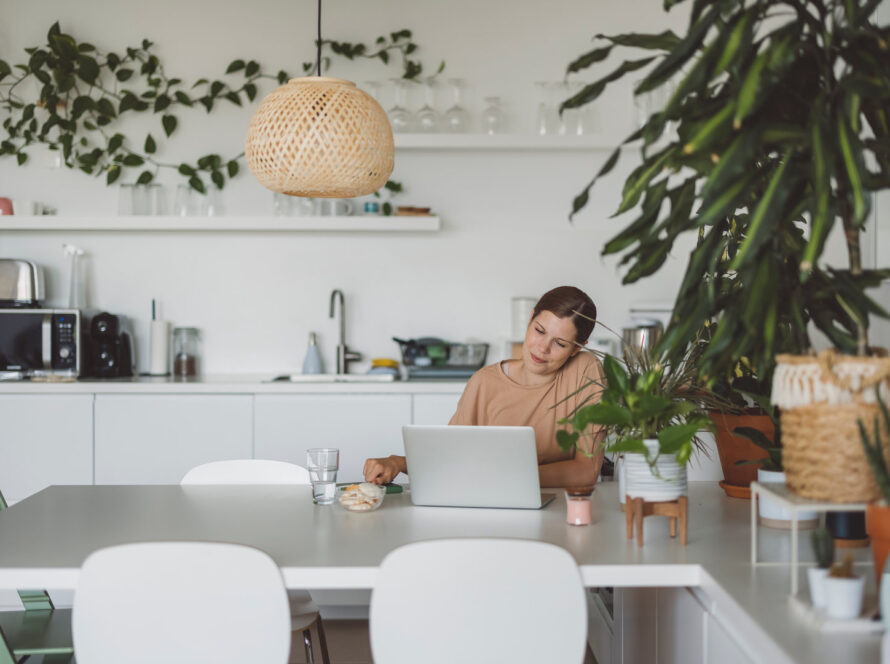 Woman in a tan top working on a laptop at a white dining table in a bright kitchen with houseplants and a rattan pendant light