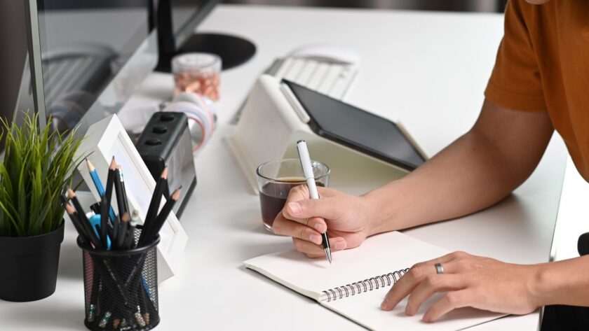Person in an orange t-shirt writing in a spiral notebook at a white desk, with a pen pot, a potted plant, a tablet and a cup of coffee beside them