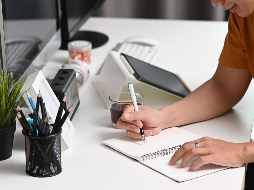 Person in an orange t-shirt writing in a spiral notebook at a white desk, with a pen pot, a potted plant, a tablet and a cup of coffee beside them